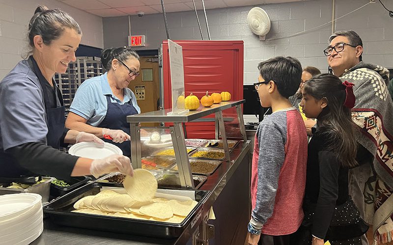 People stand waiting for their food as two women on the left serve them.