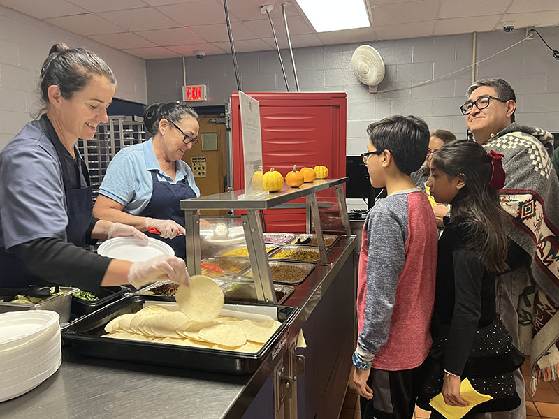 People stand waiting for their food as two women on the left serve them.