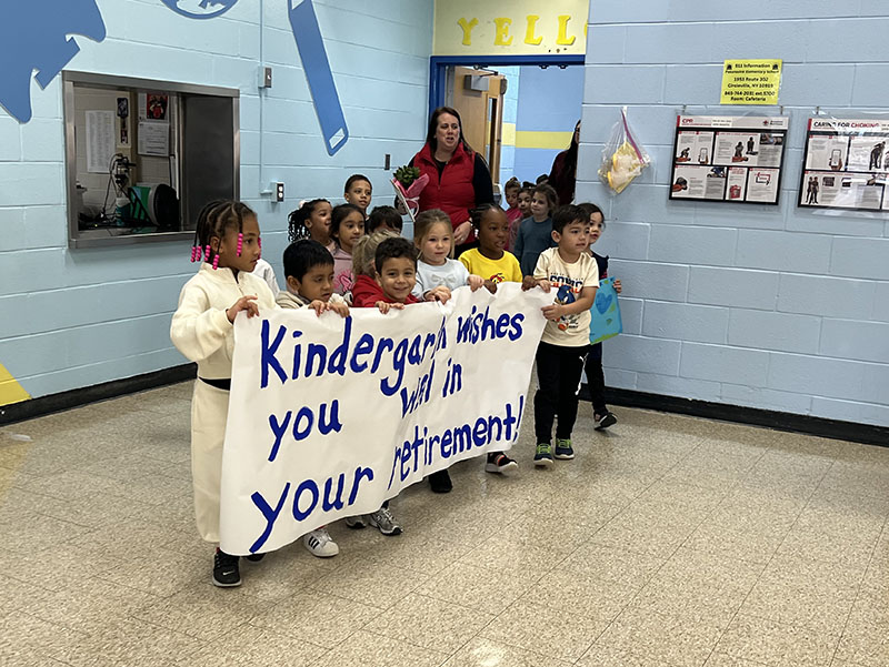 A large group of kindergarten children carry a big sign that says Kindergarten wishes you well on your retirement.