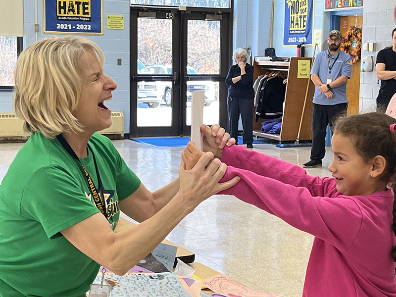 A girl in pink holds a card out in front of her, which she made, as a woman with blonde hair smiles joyfully.