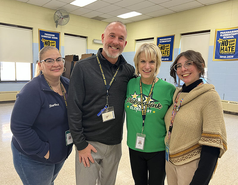 Four adults, three women and a man stand arm in arm. They are all smiling.