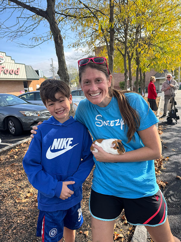 A woman with long dark hair pulled back into a ponytail smiles and holds a guinea pig. She has an arm around an elementary age boy who is also smiling.