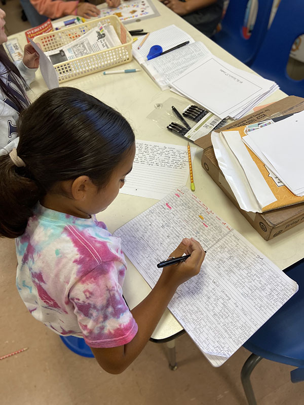 Photo from above as a fourth-grade girl writes in a notebook. The page is almost filled with writing.
