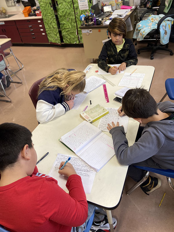 A group of four fourth-grade students sit at a long table writing in notebooks.