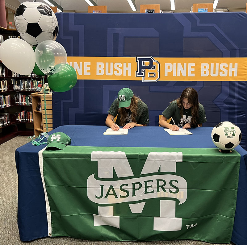 Two young women sit at a table and sign papers. The background says Pine Bush and the table cloth says Jaspers.