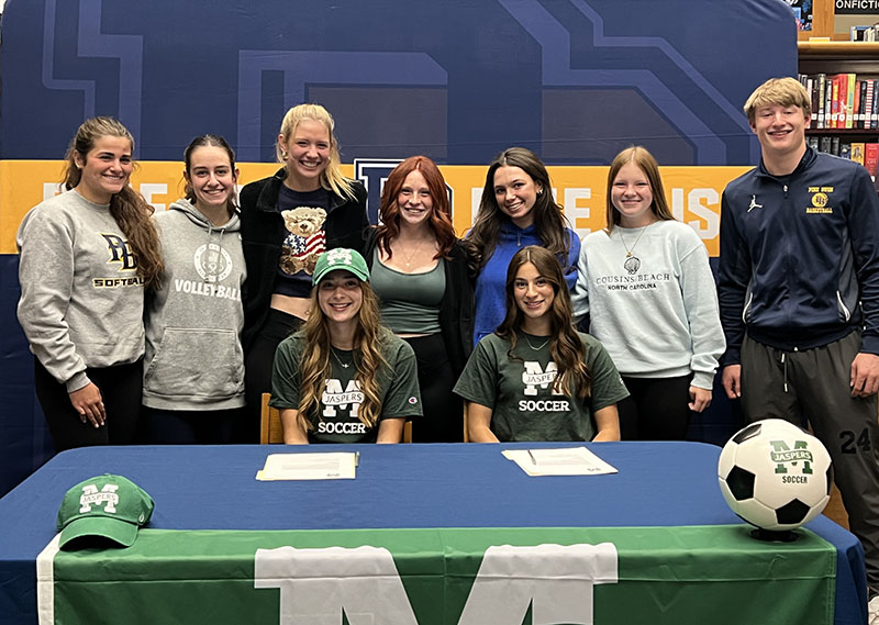 Two young women sit at a table with a group of friends behind them. All are smiling.