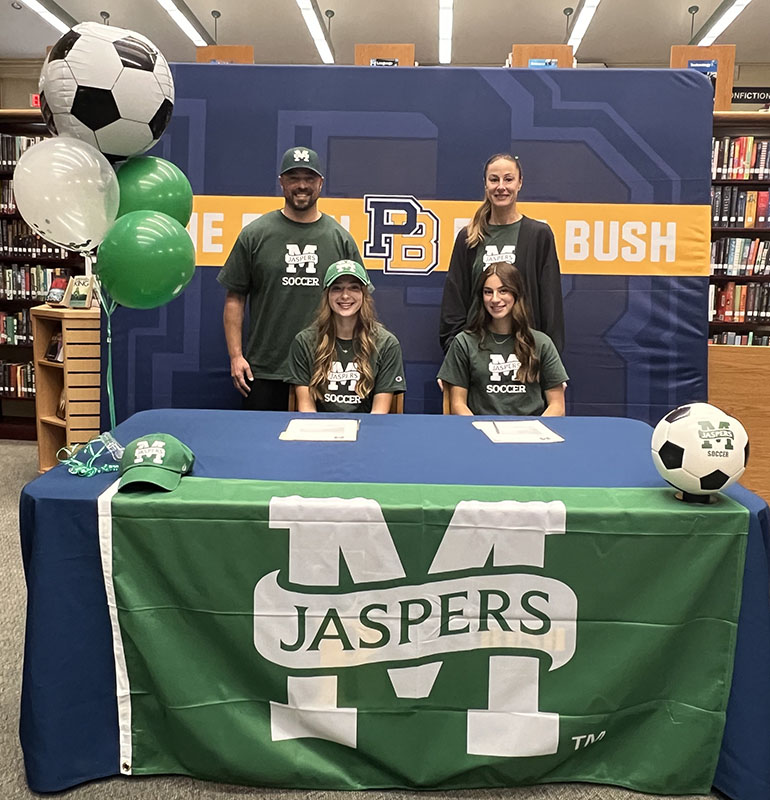 Two young women smile as they sit at a table with balloons on one side. Behind them are their parents. The banner on the front says Jaspers.