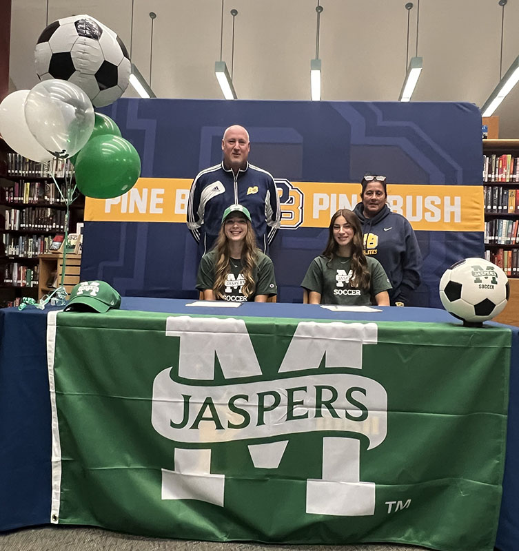 Two young women sit at a table smiling. A man and woman stand behind them. The tablecloth said Jaspers and there are green and white balloons on the side.