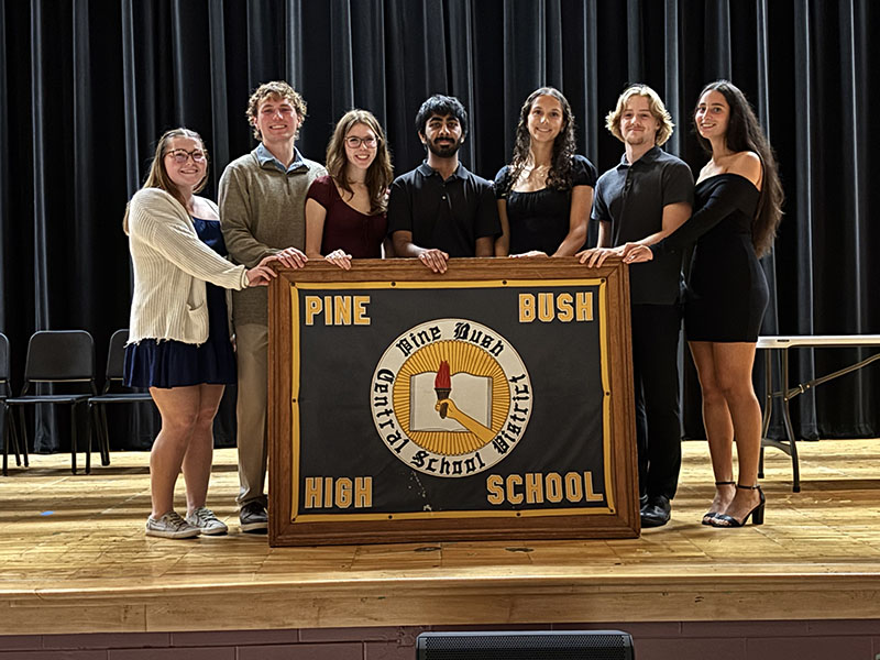 A group of seven high school students stand around a wooden podium that says Pine Bush High School. They are all dressed nicely.