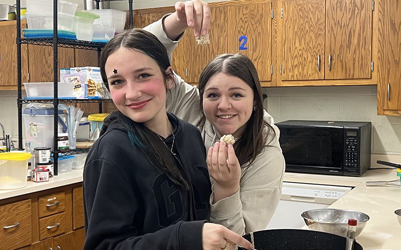 Two high school girls smile broadly. One is holding a donut they just made and the other has her spatula in the pan.