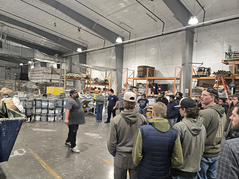 A large group of high school students stand on the floor of a warehouse listening to someone speak.