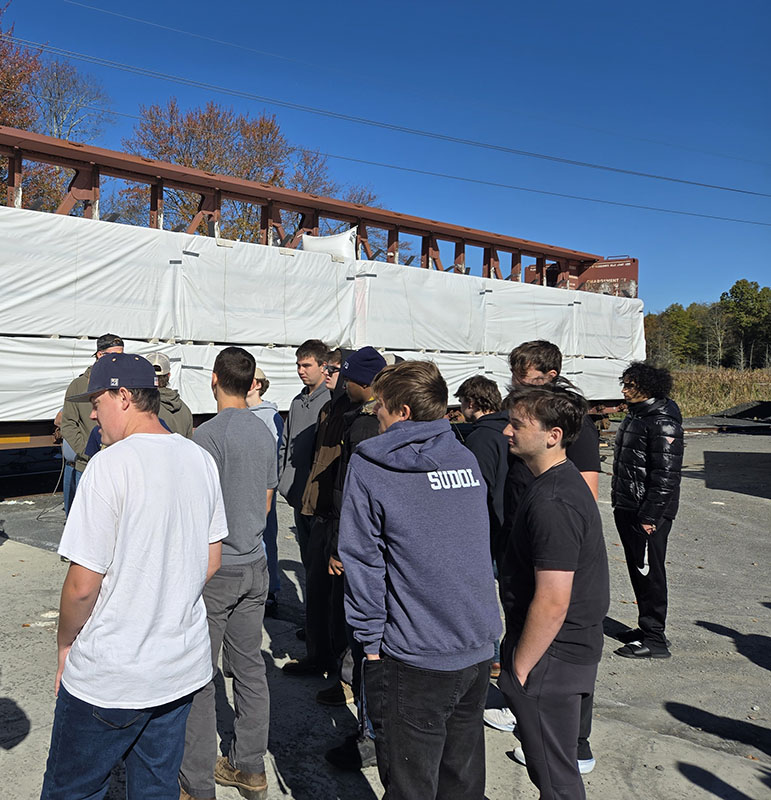 Outside, a group of high school kids stand before a large rack of wood to be used in manufacturing.