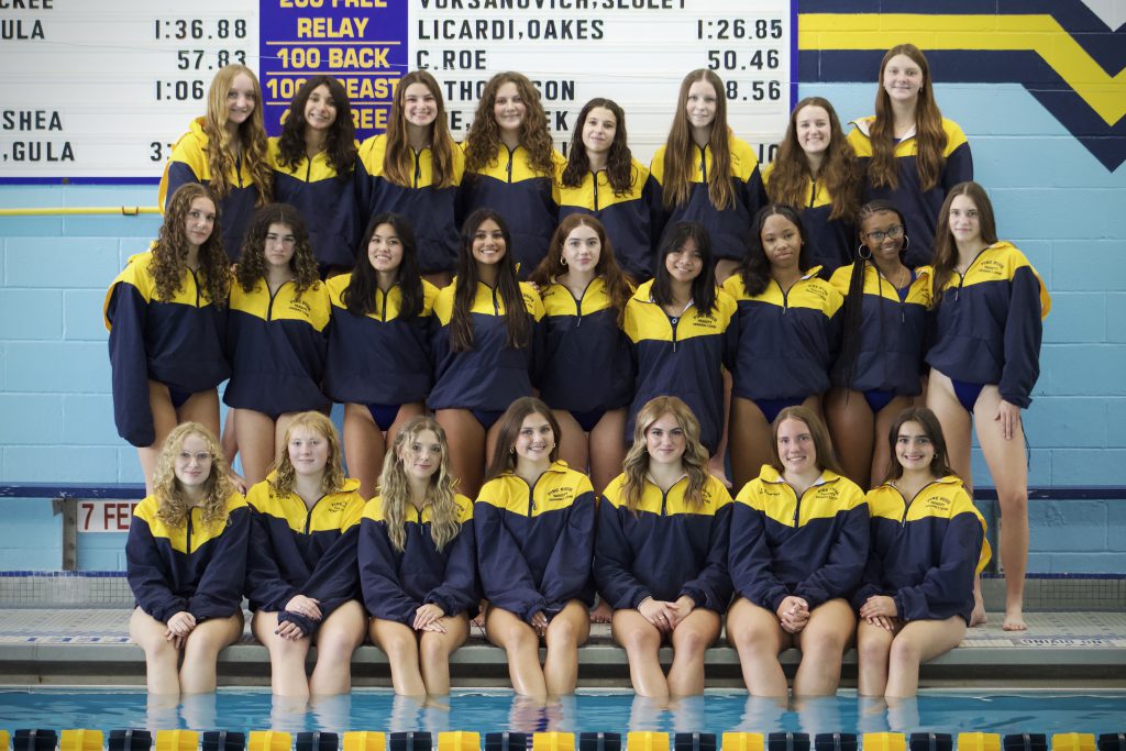 Twenty-four young women, from grades 7-12, stand in their blue and gold jackets by a pool.