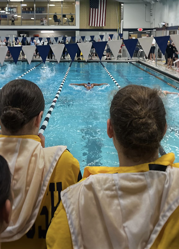 Two young women stand watching a swimmer swimming toward them.