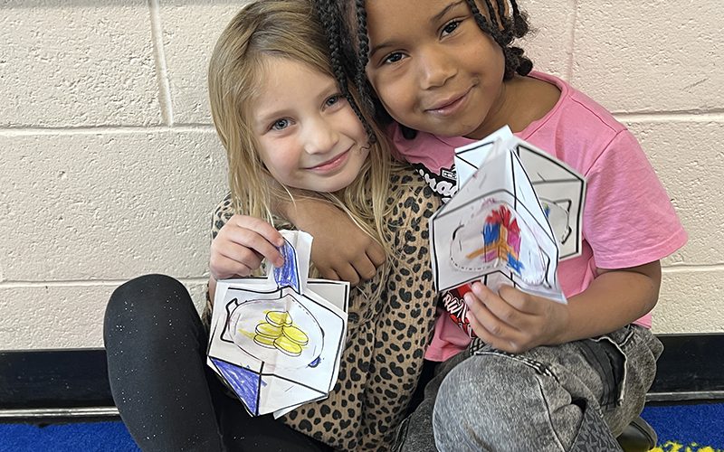 Two kindergarten kids, a girl with long blonde hair and a boy with longer dark hair, smile as they hold up their paper dreidels they made and colored.