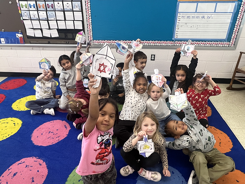 A kindergarten class sits on a colorful rug and holds up the paper dreidels they colored.