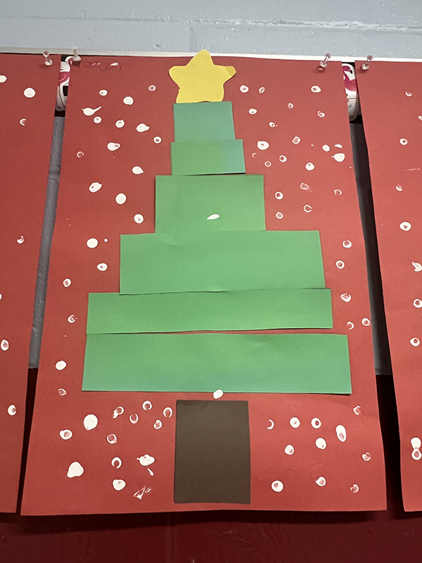 A red piece of construction paper with green strips making a Christmas tree. There are spots of white for snow and a gold star on top.