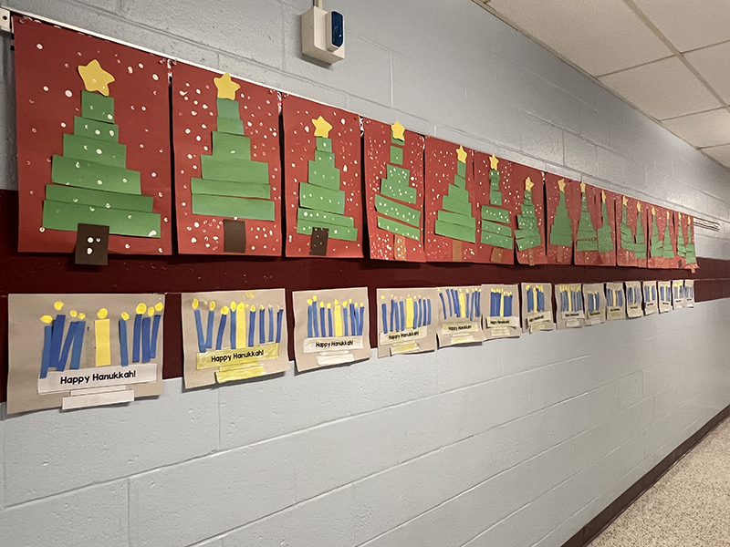 A row of menorahs and Christmas trees made on construction paper hanging in a hallway.