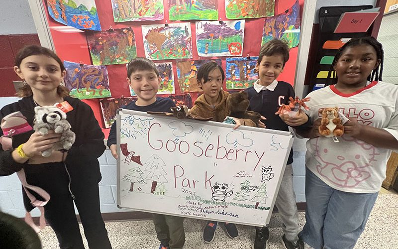 A group of five elementary age kids stand together and hold a sign that says Gooseberry Park, along with stuffed animals.