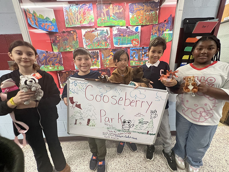 A group of five elementary age kids stand together and hold a sign that says Gooseberry Park, along with stuffed animals.