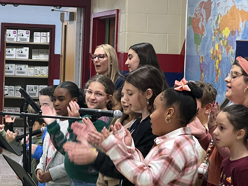 Several elementary students stand together singing. some are clapping hands.