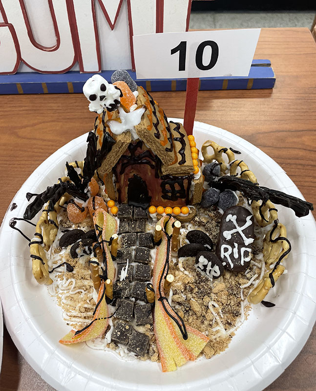 A gingerbread house made with a halloween theme with orange and black icing on the roof, a skull on the roof and cookies that look like headstones in the yard.