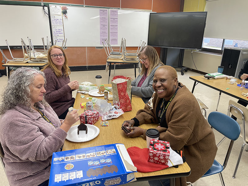 Four women sit at a table making gingerbread houses.