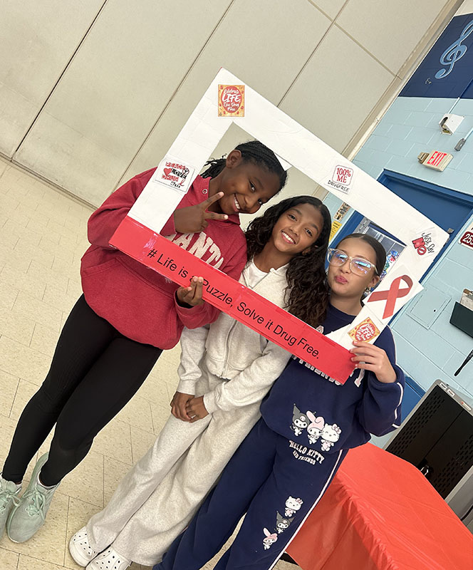Three middle school kids smile. They are holding a red and white frame that says be drug free.