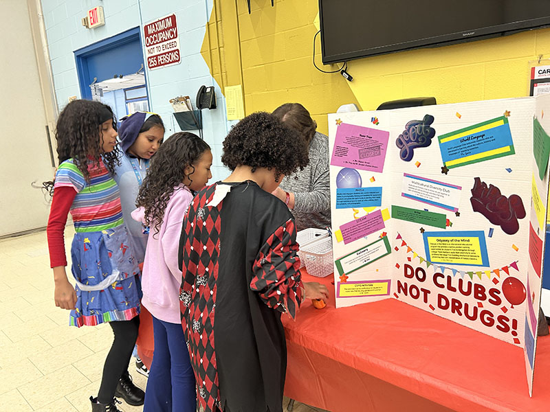 Middle school students stand at a table that has a list of clubs offered on a large white sign.