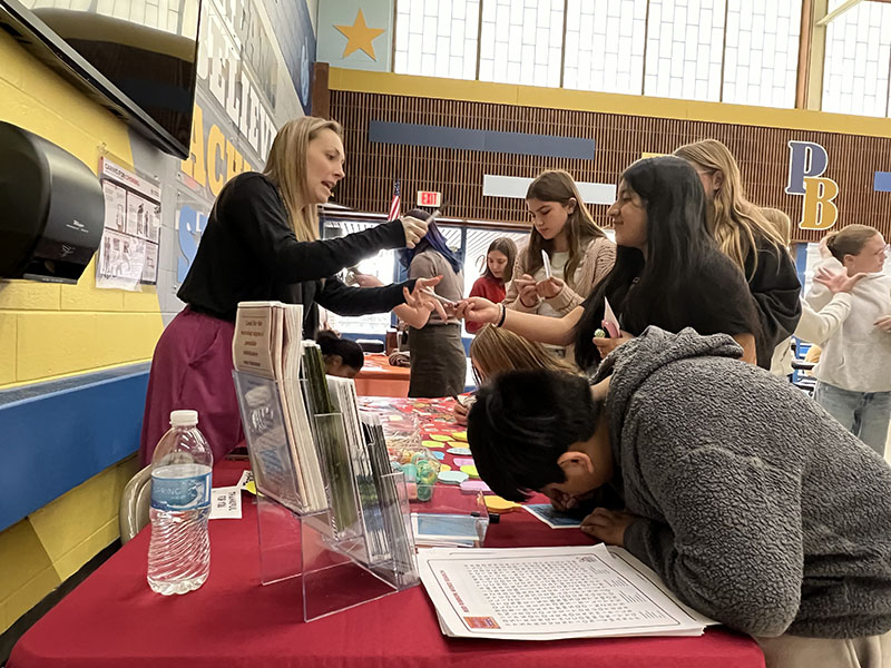 A woman in a black shirt, with long blonde hair, talks to middle schools kids at a table.