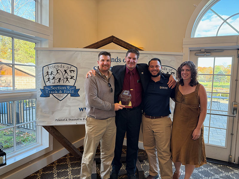 Four adults stand arm in arm and smile. Next to them is a sign that says 2025 Hall of Fame dinner Section 9 Track and Field