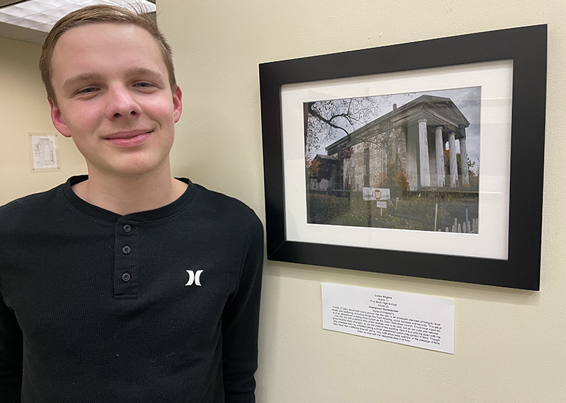 A young man stands next to a photograph hanging on the wall.