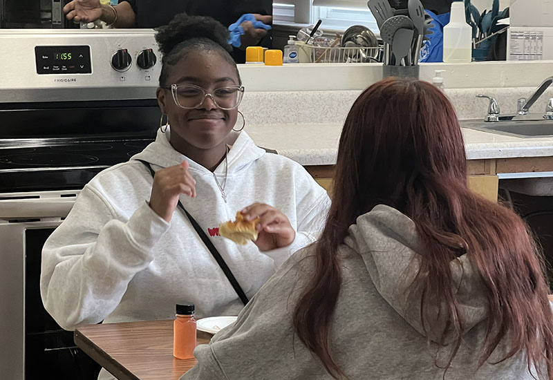 Two middle school girls sit and eat together at a table, learning about culinary arts.