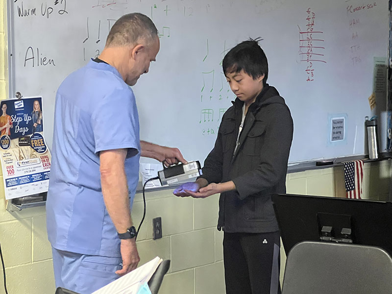 A man in scribs uses a black light to scan a middle school kid's hands.