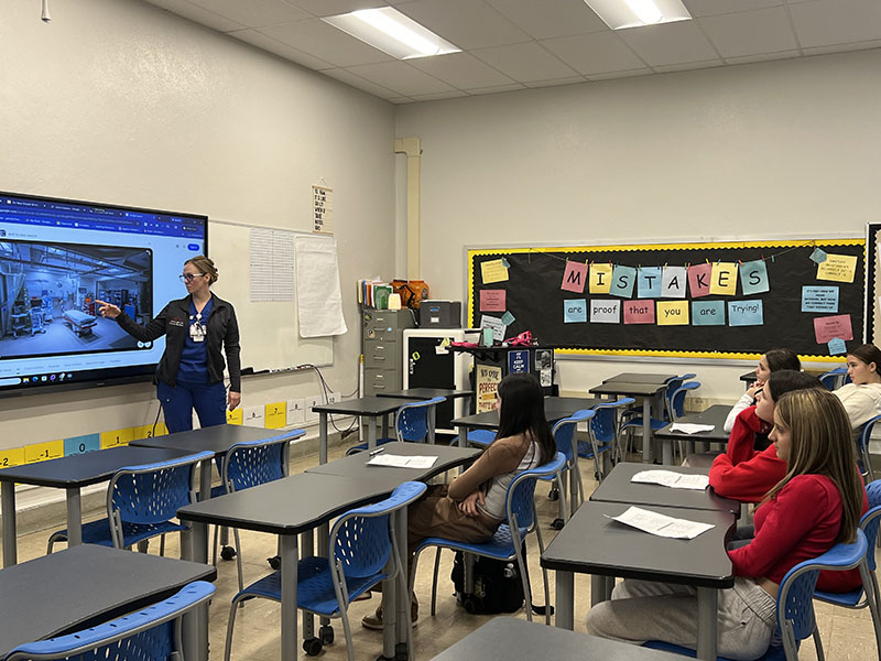 A woman stands in front of a classroom at a screen talking to students about nursing.