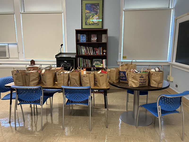 A number of brown bags filled with food sit on several desks.