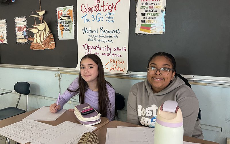 Two middle school girls sit at desks working and smile.