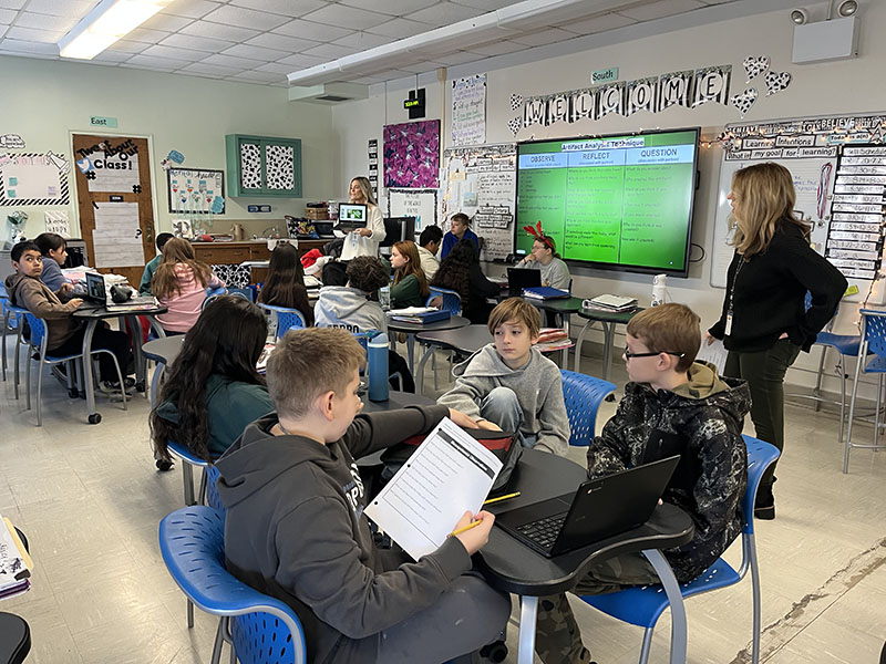 A classroom filled with middle school students look at papers and at a screen on the front wall as two teachers walk to different desks to help.