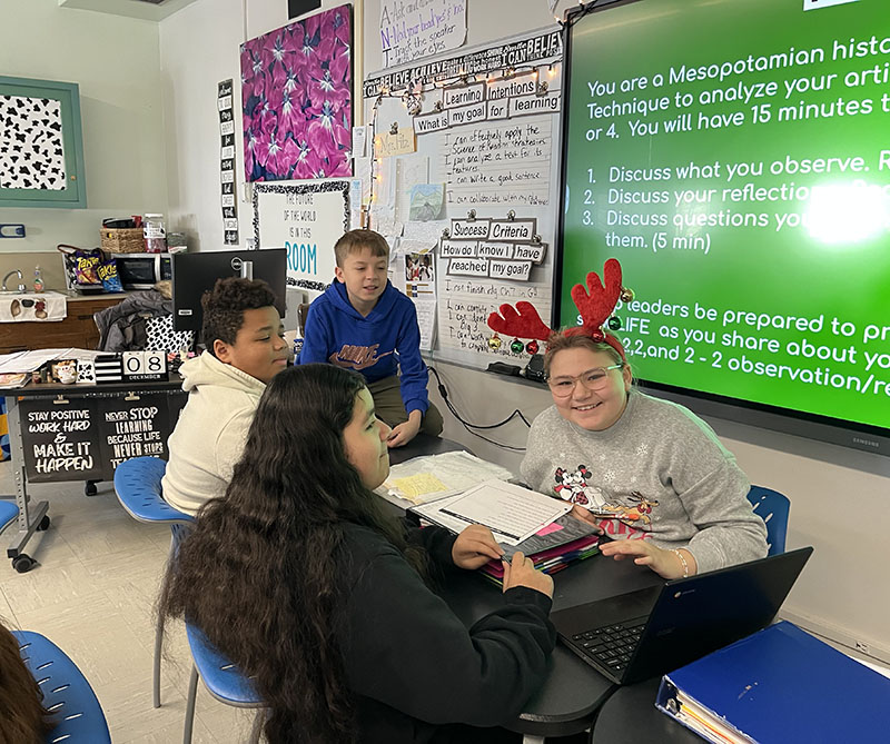Four middle school students  work in a group. There is a green board behind them with text on it.