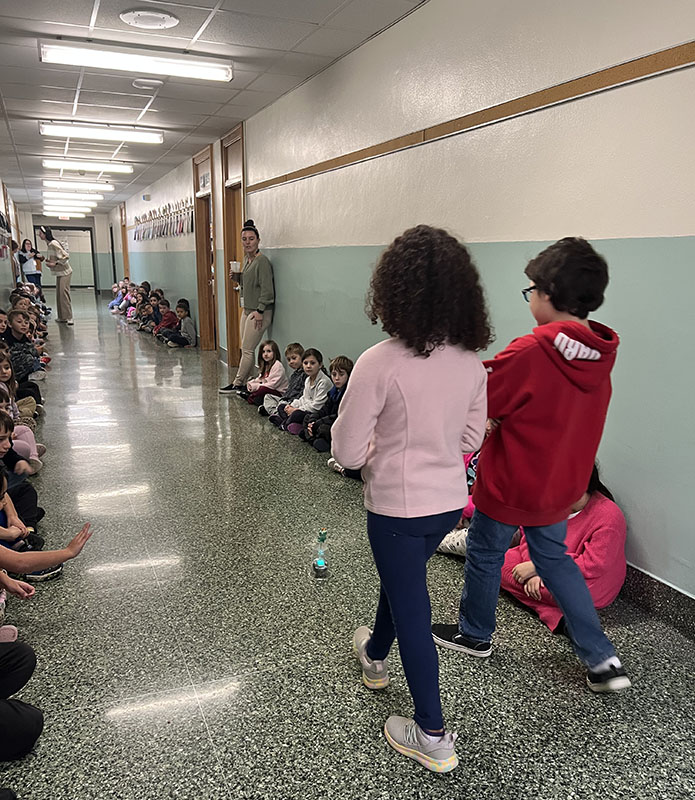 Two fourth-grade students use a remote to control a sphero robot down a hallway that is lined with younger students.