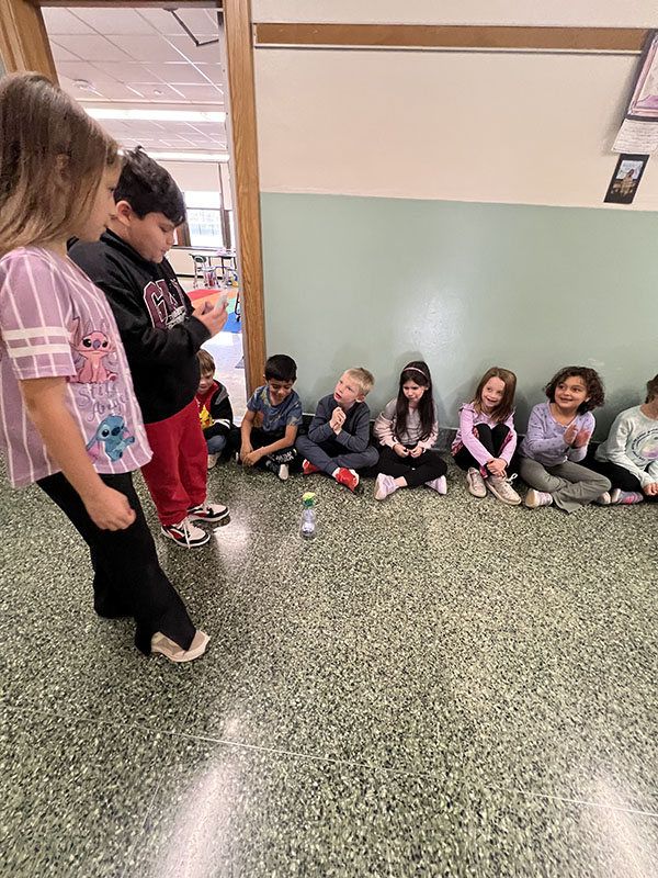 A group of smaller elementary students sit on a flor and watch as two fourth-grade students control their robot going down the hall.