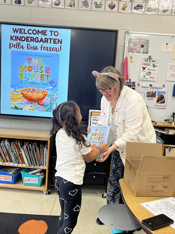 A woman hands a children's book to a little girl with long dark hair.