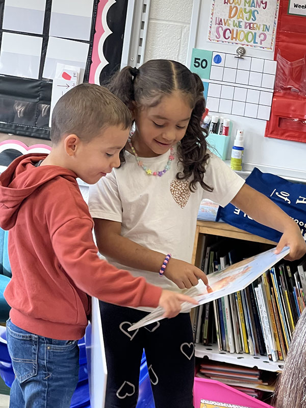 A girl in a white shirt with long dark hair stands with a boy in a red sweatshirt and short hair. They are looking together in a book and pointing to things.