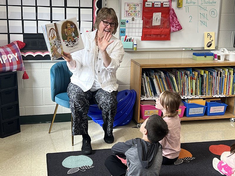 A woman with glasses and shorter light hair sits in a chair holding a book open and reads to little kids sitting on a carpet.
