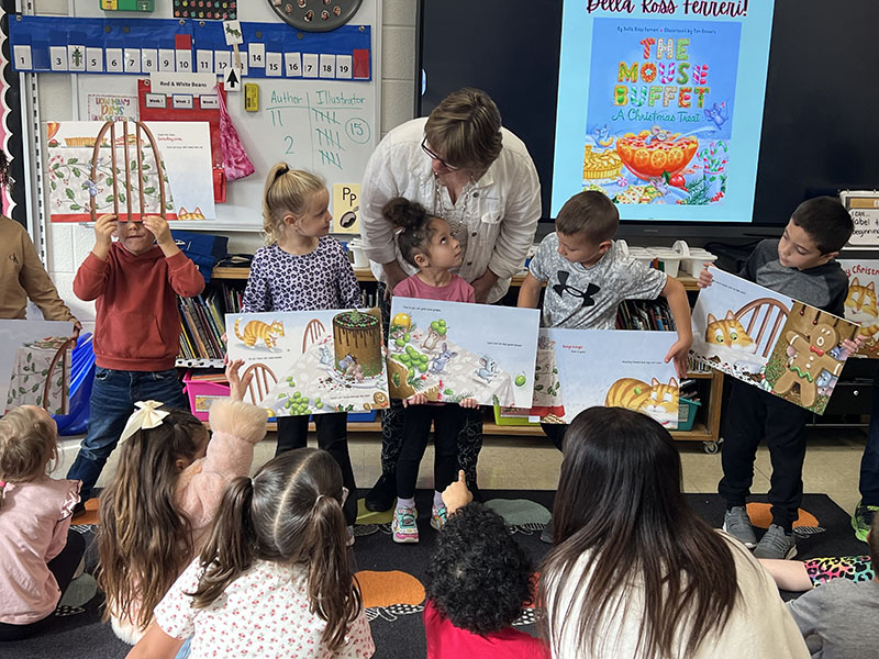 Kindergarten children hold story boards from a book. They are in front of a group of their classmates.