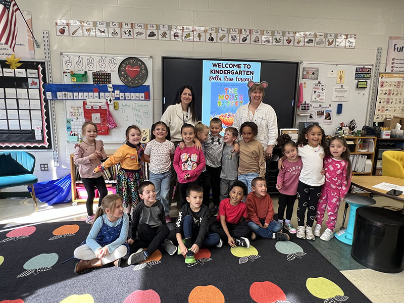 A full class of kindergarten kids stand with two adults. Some are holding books.