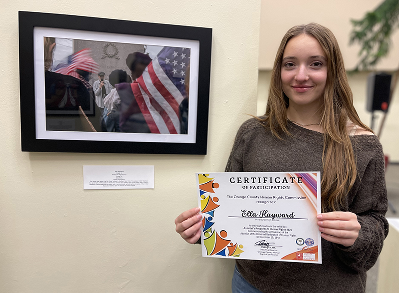 A young woman smiles and holds a certificate. She is standing next to a photograph hanging on a wall.