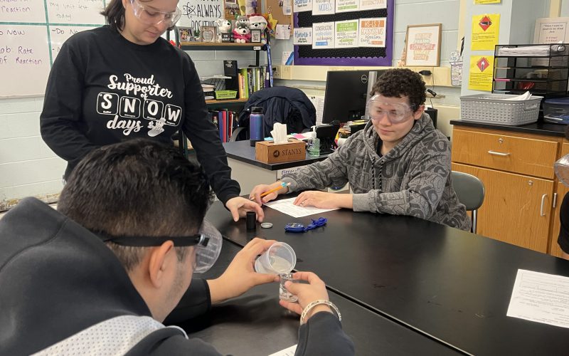 Middle school students sit at tables in a lab. They re wearing goggles. One is measuring a liquid. A teacher stands by them.