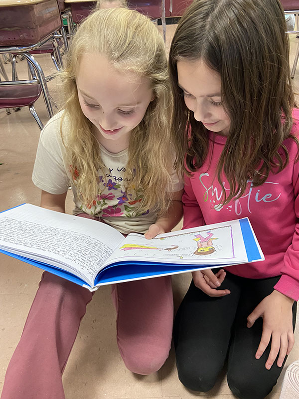 Two fourth-grade girls smile as they look at a book they are holding opened in front of them.