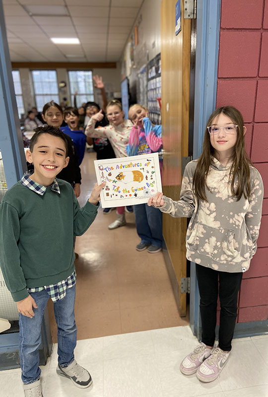 Standing in a doorway, two fourth grade kids hold a book. Behind them are their classmates smiling.